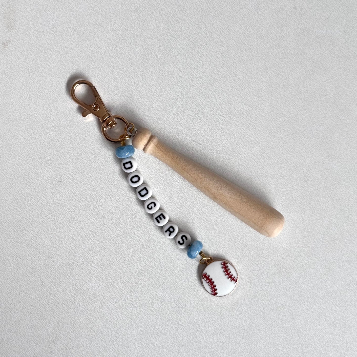 A baseball-themed keychain featuring a light wood baseball bat, a gold-tone lobster clasp, white letter beads spelling 'DODGERS', light blue beads, and a small baseball charm on a light gray background.